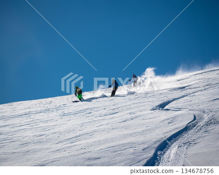 Three skiers carve down a sunny, powder-covered mountain slope, kicking up fresh snow under a clear blue sky, creating a dynamic winter sports action scene Three skiers carve down a sunny, powder-covered mountain slope, kicking up fresh snow under a clear blue sky, creating a dynamic winter sports action scene 134678756