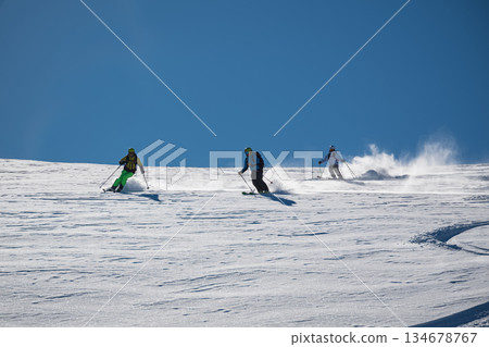 Three skiers carve down a sunny, powder-covered mountain slope, kicking up fresh snow under a clear blue sky, creating a dynamic winter sports action scene Three skiers carve down a sunny, powder-covered mountain slope, kicking up fresh snow under a clear blue sky, creating a dynamic winter sports action scene 134678767