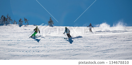 Three skiers carve down a sunny, powder-covered mountain slope, kicking up fresh snow under a clear blue sky, creating a dynamic winter sports action scene 134678810