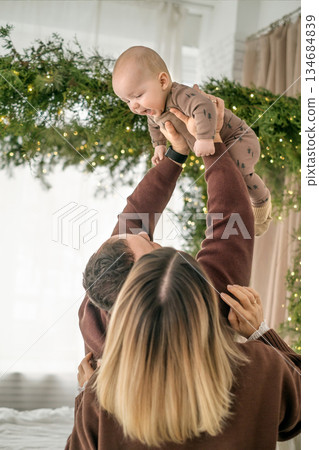 A father plays with baby son, lifting him high above himself. parents play with their children in a bedroom decorated for Christmas and New Year. 134684839