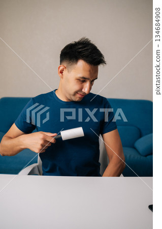 Vertical portrait of smiling man sitting at white table using sticky lint roller to remove dust, hair and lint from blue t-shirt, looking pleased with cleaning results in modern apartment. Vertical portrait of smiling man sitting at white table using sticky lint roller to remove dust, hair and lint from blue t-shirt, looking pleased with cleaning results in modern apartment. 134684908