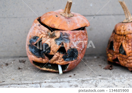 Rotting Halloween pumpkin with a carved face on the ground with a cigarette. Rotting Halloween pumpkin with a carved face on the ground with a cigarette. 134685900