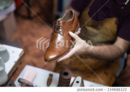 Cobbler presents a polished brown leather wingtip dress shoe in a craftsman's workshop Cobbler presents a polished brown leather wingtip dress shoe in a craftsman's workshop 134686122
