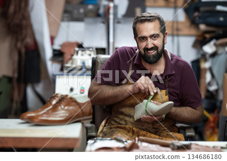Smiling shoemaker at work in leather workshop measuring a shoe with a tape, showcasing handmade craftsmanship Smiling shoemaker at work in leather workshop measuring a shoe with a tape, showcasing handmade craftsmanship 134686180