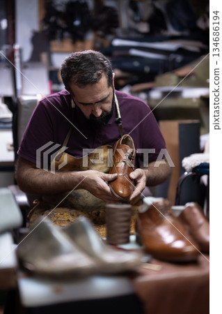 Cobbler expertly crafts a brown leather shoe in a bustling workshop environment 134686194