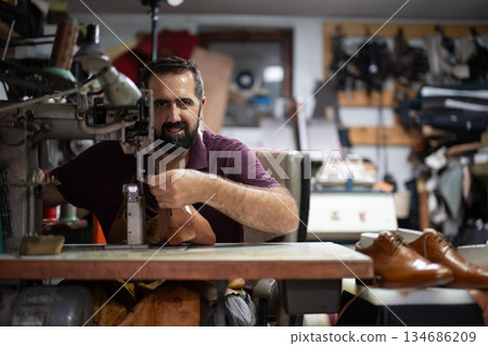 Skilled craftsman operating a sewing machine in a cluttered leather workshop making shoes Skilled craftsman operating a sewing machine in a cluttered leather workshop making shoes 134686209