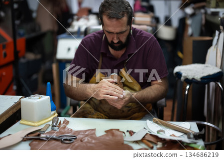 Leather artisan shapes leather at a cluttered workshop bench, focused on handcrafting a piece Leather artisan shapes leather at a cluttered workshop bench, focused on handcrafting a piece 134686219