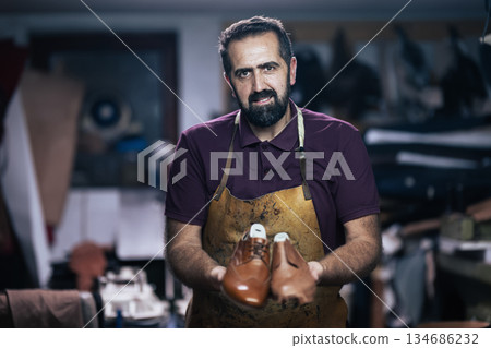 Skilled craftsman in a workshop presenting brown leather shoes, wearing an apron and focused on his craft Skilled craftsman in a workshop presenting brown leather shoes, wearing an apron and focused on his craft 134686232