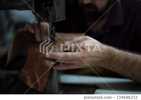 Craftsman guides brown leather under a sewing machine, stitching a piece with careful, precise handiwork. Craftsman guides brown leather under a sewing machine, stitching a piece with careful, precise handiwork. 134686252