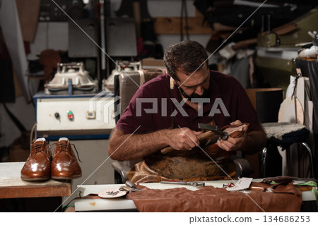 Leather shoemaker at work in a busy workshop shaping leather, with brown leather shoes on the table nearby Leather shoemaker at work in a busy workshop shaping leather, with brown leather shoes on the table nearby 134686253