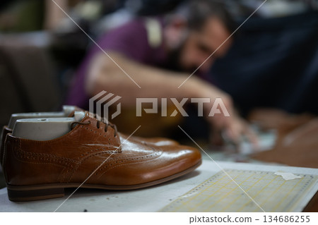 Brown leather dress shoe on a workbench with a cobbler at work in the background Brown leather dress shoe on a workbench with a cobbler at work in the background 134686255