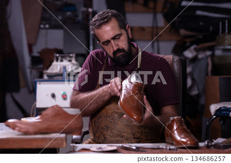 Cobbler crafts brown leather shoes in a workshop, focusing on detailing and shaping Cobbler crafts brown leather shoes in a workshop, focusing on detailing and shaping 134686257