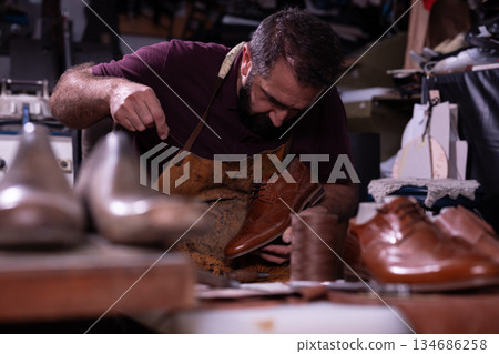 Skilled craftsman handcrafts brown leather shoe in a busy workshop filled with tools 134686258