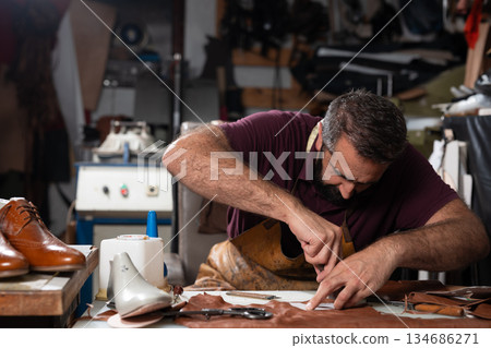 Man crafts leather shoes in a cluttered workshop, focused on stitching and detailing 134686271