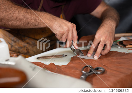 Leather craft in progress: close-up of hands cutting leather with a knife and tools Leather craft in progress: close-up of hands cutting leather with a knife and tools 134686285