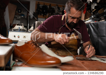 Shoemaker at work crafting leather shoes with hammer and chisel in a traditional workshop Shoemaker at work crafting leather shoes with hammer and chisel in a traditional workshop 134686290