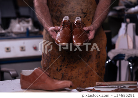 Cobbler holds polished brown leather dress shoes in workshop with tools on the workbench Cobbler holds polished brown leather dress shoes in workshop with tools on the workbench 134686291