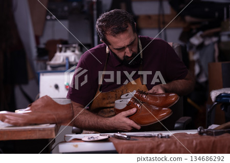 Shoemaker carefully inspects brown leather dress shoes in a busy workshop, highlighting craftsmanship and traditional handwork Shoemaker carefully inspects brown leather dress shoes in a busy workshop, highlighting craftsmanship and traditional handwork 134686292