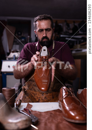 Cobbler presents a polished brown leather oxford shoe in a busy workshop with tools 134686293
