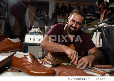 Smiling shoemaker at a leather workshop crafting brown leather shoes with tools and stitching 134686299