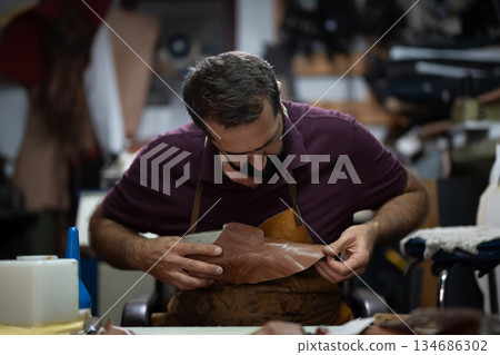 Cobbler shaping leather shoe in a busy workshop, a focused artisan at work on intricate stitching Cobbler shaping leather shoe in a busy workshop, a focused artisan at work on intricate stitching 134686302