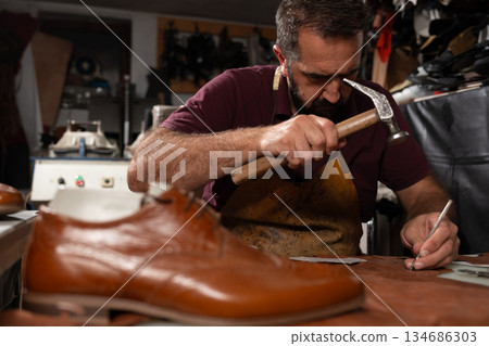 Cobbler at work repairing leather shoe with hammer and awl in a focused old-world workshop Cobbler at work repairing leather shoe with hammer and awl in a focused old-world workshop 134686303