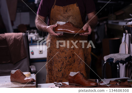 Cobbler craftsman holds a brown leather shoe in a busy workshop full of tools and other footwear 134686308