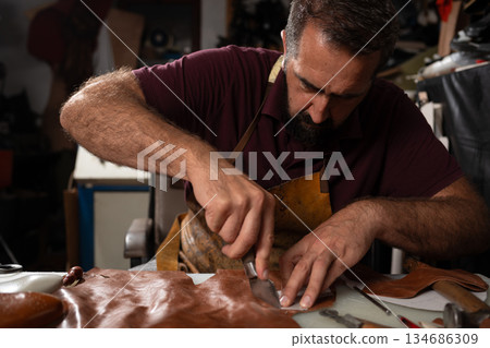 A skilled craftsman carves and cuts leather in a workshop, shaping a belt at the studio. A skilled craftsman carves and cuts leather in a workshop, shaping a belt at the studio. 134686309