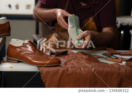 Skilled artisan examines a leather sole with a measuring tape in a busy shoemaking workshop 134686322
