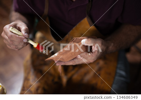 Artisan hand paints leather piece with brush, crafting detailed leatherwork by hand Artisan hand paints leather piece with brush, crafting detailed leatherwork by hand 134686349