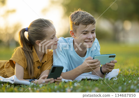 Positive children boy and girl playing online games on their mobile phones with friends outdoors in summer park. Positive children boy and girl playing online games on their mobile phones with friends outdoors in summer park. 134687850