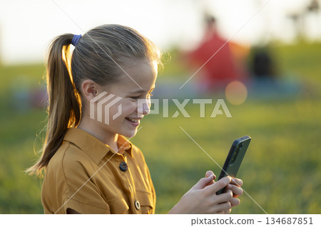 Positive child girl playing online game on her mobile phone with a friend outdoors in summer park 134687851