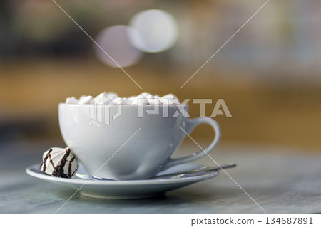 Mug of coffee with marshmallows on porcelain plate on blurred colorful interior bokeh background. 134687891
