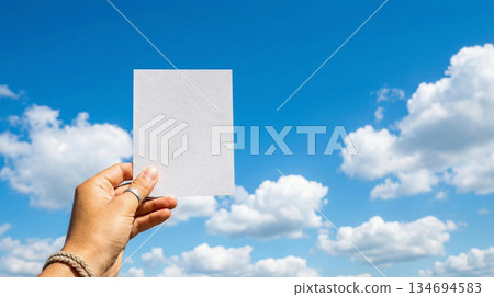 A hand (faceless) holding a flyer up against a blue sky with fluffy white clouds 134694583