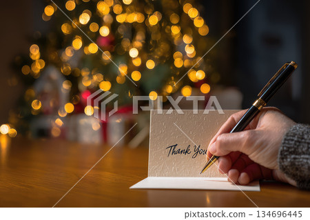 Writing thank you notes during festive season brings warmth and gratitude with glowing holiday background and hand holding pen on wooden table 134696445