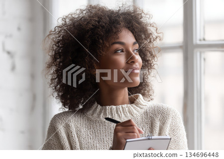 Writing in five minute journal, young woman with curly hair and cozy sweater sits by bright window, smiling thoughtfully as she reflects and plans her day 134696448