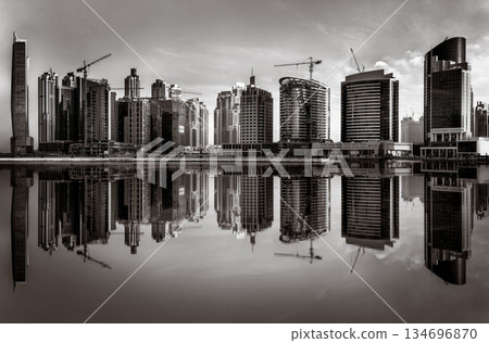 Monochrome city skyline with tall skyscrapers reflecting on calm water under a cloudy sky, Dubai Marina bay UAE 134696870