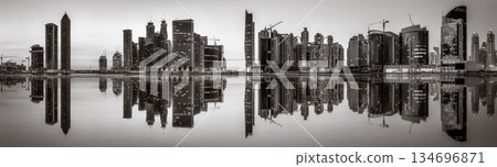 Monochrome city skyline with tall skyscrapers reflecting on calm water under a cloudy sky, Dubai Marina bay UAE Monochrome city skyline with tall skyscrapers reflecting on calm water under a cloudy sky, Dubai Marina bay UAE 134696871