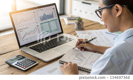 Woman reviews financial data on a laptop, taking notes in a 134697368