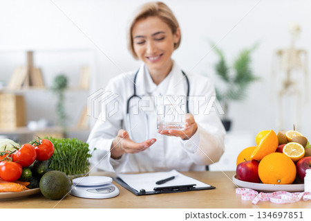A smiling dietitian offers a glass of water, surrounded by fresh fruits and vegetables, promoting healthy eating habits. 134697851