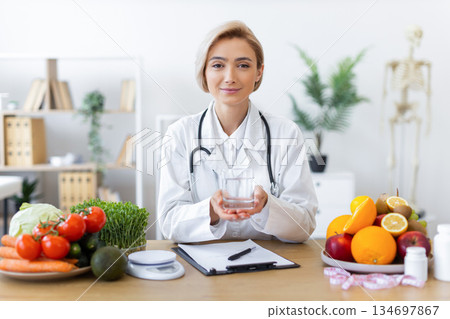 A nutritionist holds a glass of water, surrounded by fresh fruits and vegetables, promoting healthy eating habits. A nutritionist holds a glass of water, surrounded by fresh fruits and vegetables, promoting healthy eating habits. 134697867