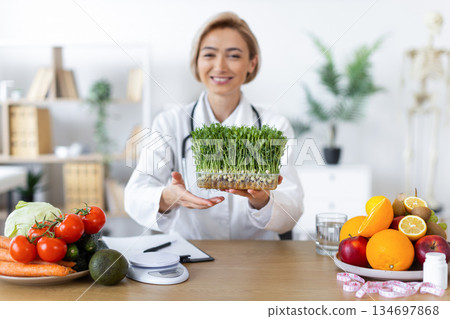 A smiling nutritionist presents a tray of microgreens, surrounded by fresh fruits and vegetables, promoting healthy eating. A smiling nutritionist presents a tray of microgreens, surrounded by fresh fruits and vegetables, promoting healthy eating. 134697868
