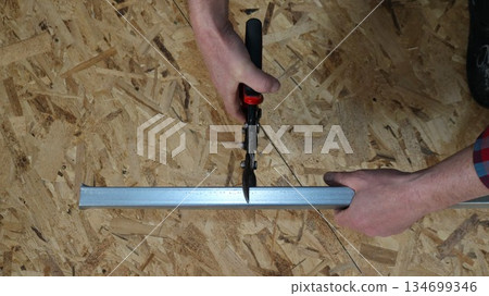 Hands holding a narrow metal profile and metal scissors with a long handle, top view against the background of a textured light OSB sheet, cutting metal frame material with a special tool 134699346