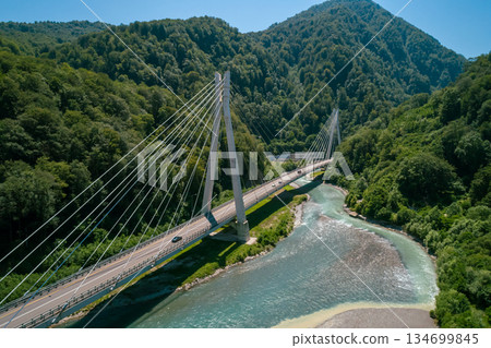 A cable-stayed bridge spans a river surrounded by lush green mountains, with cars traveling along the road A cable-stayed bridge spans a river surrounded by lush green mountains, with cars traveling along the road 134699845