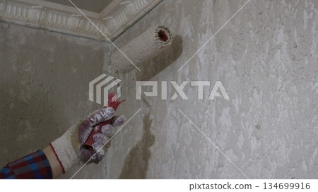 The left hand in a glove holds a roller and applies mortar, putty, primer or paint to the wall under the ceiling, repair work in the room, preparing interior walls for renovation The left hand in a glove holds a roller and applies mortar, putty, primer or paint to the wall under the ceiling, repair work in the room, preparing interior walls for renovation 134699916