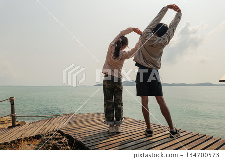 Travel couple enjoying holiday happy relax during nature adventure, stretching bodies together on wooden pier, pristine ocean water and vast sky creating serene outdoor scene Travel couple enjoying holiday happy relax during nature adventure, stretching bodies together on wooden pier, pristine ocean water and vast sky creating serene outdoor scene 134700573