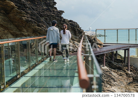 Happy travel couple enjoys holiday adventure, relaxing on glass skywalk beside rugged nature cliff, exploring beautiful coastal path with clear calm ocean view 134700578