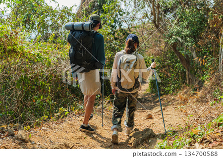 Travel young couple hike mountain forest path, people enjoy walk autumn nature journey, carrying backpack and trekking pole, exploring green foliage on sunny day for recreation 134700588