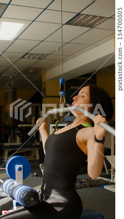 Vertical photo of a Caucasian woman in excellent physical shape during gym working out. Workout for back muscles. Physical education as a norm of life for maintaining health. 134700805