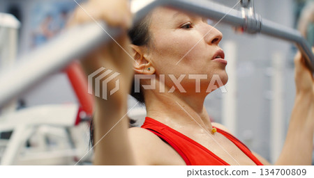 Close-up of a fitness woman working out pulldown training at gym. A woman doing exercise on a lat machine in the gym. A woman performs lat pulldowns to strengthen and tone her upper back muscles. Close-up of a fitness woman working out pulldown training at gym. A woman doing exercise on a lat machine in the gym. A woman performs lat pulldowns to strengthen and tone her upper back muscles. 134700809